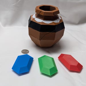 May include: Three plastic gems, one blue, one green, and one red, are arranged on a white surface. A brown, segmented, ceramic pot sits in the background.