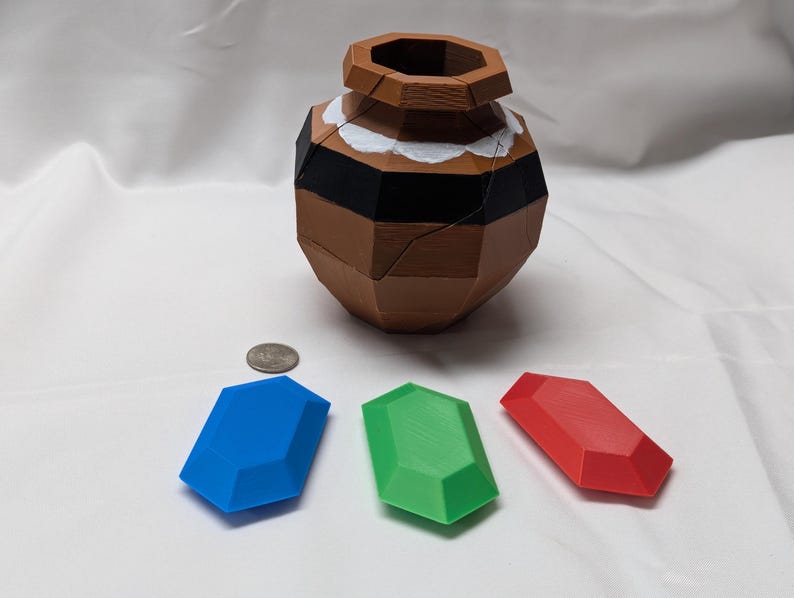May include: Three plastic gems, one blue, one green, and one red, are arranged on a white surface. A brown, segmented, ceramic pot sits in the background.