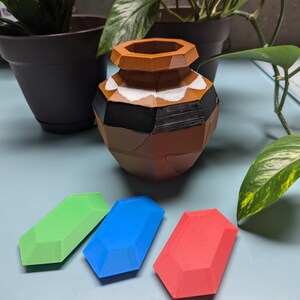 May include: Three 3D printed gems in green, blue, and red, sitting on a light blue surface. A brown and white ceramic pot is in the background.