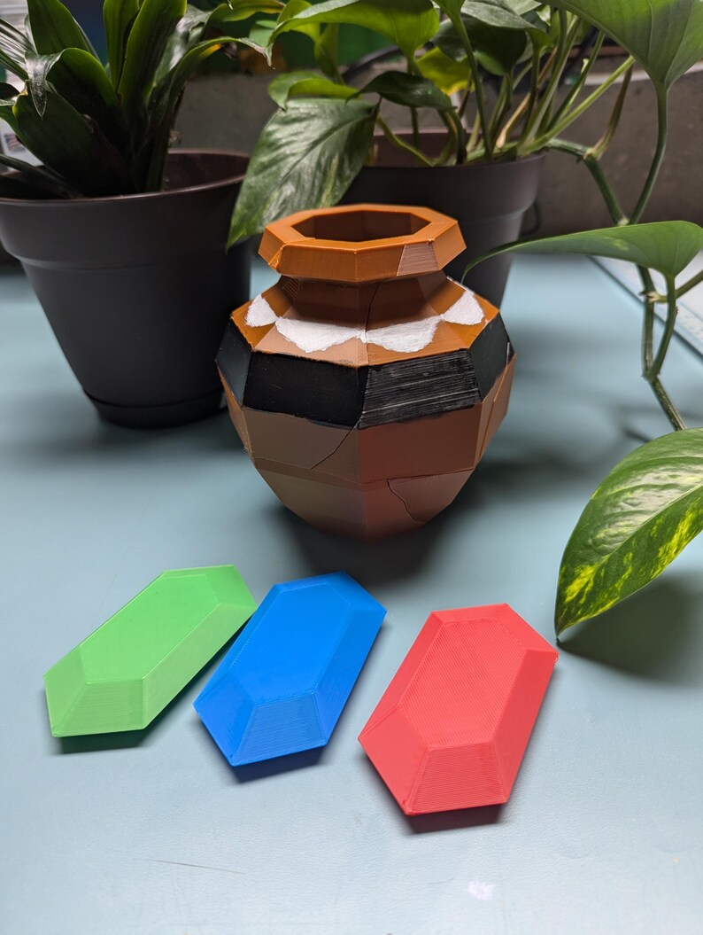 May include: Three 3D printed gems in green, blue, and red, sitting on a light blue surface. A brown and white ceramic pot is in the background.