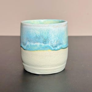 Könnte beinhalten: Ein Keramikbecher mit zweifarbigem Design, bestehend aus einer glatten, matten cremeweißen Basis und einer glänzenden, strukturierten türkisfarbenen Glasur, die vom Rand herabläuft. Das Gefäß hat eine zylindrische Form mit leicht abgerundeten Seiten.