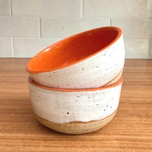 May include: Two stacked ceramic bowls featuring a bright orange interior and a white exterior with dark speckles. The base of the bottom bowl reveals a natural tan clay finish. The bowls have a rounded shape and a visible hand-crafted texture.