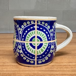 Puede incluir: Una taza de cerámica con asa y borde blancos. La taza presenta un fondo azul con un diseño geométrico en verde y blanco. El diseño incluye círculos, líneas y un patrón en forma de cruz.