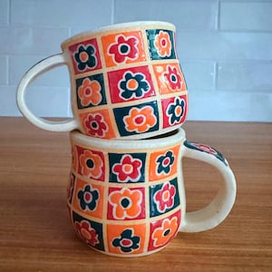 May include: Two ceramic mugs stacked vertically, each featuring a grid pattern of alternating squares. The squares contain stylized flower motifs in vibrant shades of orange, red, and dark green against a cream-colored background.