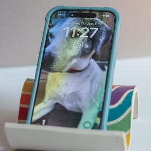 May include: A teal smartphone in a teal case displays a dog image on the screen, propped on a white and rainbow-colored phone stand. The screen shows "Sunday, October 5" and the time "11:27". The phone stand has a curved design.