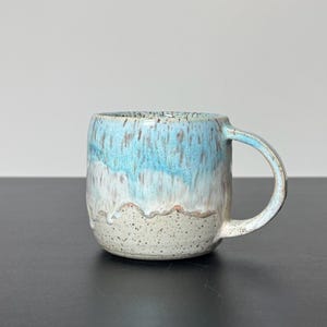 May include: A ceramic mug with a rounded shape and a curved handle. The upper portion features a glossy, drippy light blue glaze with brown speckles, while the lower section is a matte, speckled off-white clay with a textured, uneven finish.