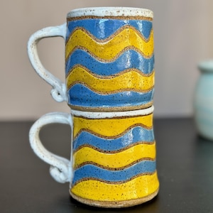 Könnte beinhalten: Zwei handgefertigte Keramikbecher übereinander gestapelt. Jeder Becher hat ein wellenförmiges Muster aus blauen und gelben Streifen. Die Griffe und Ränder sind weiß. Die Becher sind etwa 10 cm hoch.