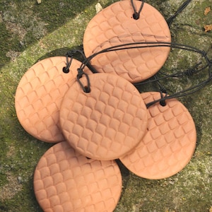 May include: Several terracotta-coloured, circular pendants with a textured, diamond-patterned surface. Each pendant is strung on a black cord. The pendants are arranged on a mossy, green surface.