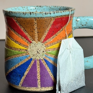 May include: A ceramic mug with a turquoise handle and rim. The mug features a colorful sunburst design in shades of red, orange, yellow, green, blue, and purple. A tea bag hangs from the rim of the mug.