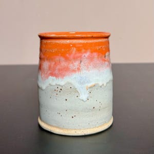 Op de afbeelding: Een cilindervormige keramische beker met een feloranje glazuur aan de bovenkant dat overgaat in een zacht wit druippatroon over een gespikkelde lichtgrijze basis. Het vat heeft een gladde rand en een platte bodem, wat een ambachtelijke uitstraling geeft.