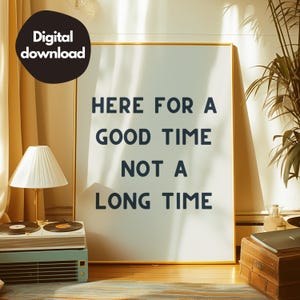Puede incluir: Una impresión enmarcada en dorado con el texto "HERE FOR A GOOD TIME NOT A LONG TIME" sobre un fondo blanco. La impresión está colgada en una pared en una habitación con una lámpara y una mesa de madera.
