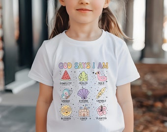 May include: White t-shirt with the text "God Says I Am" in rainbow letters. The shirt features a variety of colorful illustrations, including a watermelon, a leaf, a flamingo, a butterfly, a seashell, a surfboard, a sun, a castle, and a hibiscus flower. Each illustration is accompanied by a word and a Bible verse.