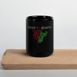 May include: Black coffee mug with the text "KEEP IT SIMPLE" in white. The mug features a geometric design of a bull and a bear in red and green. The mug is sitting on a wooden surface.