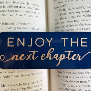 May include: A blue bookmark with silver lettering that reads "ENJOY THE next chapter". The bookmark is placed on an open book.