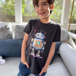 May include: A young person wearing a dark grey t-shirt with a cartoon robot graphic. The robot is blue, yellow, and orange. The person is wearing blue jeans and is kneeling on a white couch.