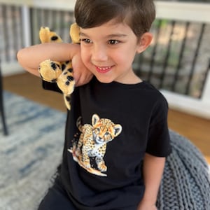 May include: A young boy wearing a black t-shirt with a cheetah print design. He is holding a stuffed cheetah toy and sitting on a gray knitted pouf.