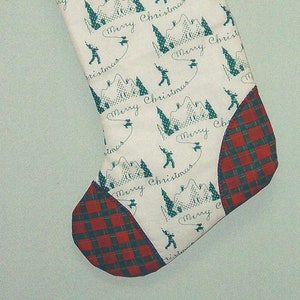 Pode incluir: Meia de Natal com punho, calcanhar e biqueira xadrez vermelhos e verdes. O corpo principal é branco com um padrão repetido de esquiadores, montanhas e as palavras "Merry Christmas" em verde-azulado.