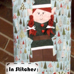 May include: A Christmas stocking with a blue and white background featuring a pattern of small Christmas trees. The stocking has a green elf applique with red hair, white bows, and red buttons. The stocking has a white label with the text "In Stitches .pdf PATTERN".