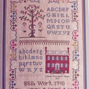 May include: A cross-stitch sampler featuring a colourful alphabet, a tree with two figures, and a red brick house. The sampler is framed and includes the text "Isaiah Davenport House 1820" and "SSB's Work 1990".