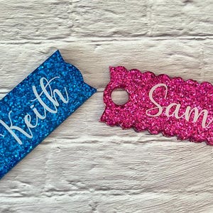 May include: Two glitter name tags. One is blue with the name "Keith" in white script, the other is pink with "Sam" in white script. Both have a scalloped edge and a hole for attachment.