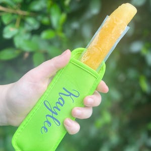 Puede incluir: Un porta-paletas verde con el nombre "Koylee" bordado en él. El porta-paletas está hecho de un material suave y aislante diseñado para mantener las paletas frías y evitar que se derritan. Una paleta amarilla está dentro del porta-paletas.