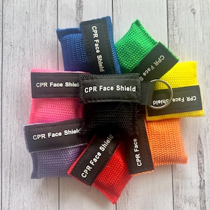 May include: A set of seven CPR face shields in various colors, including pink, purple, blue, black, green, yellow, and red. Each shield has the text "CPR Face Shield" printed on it.