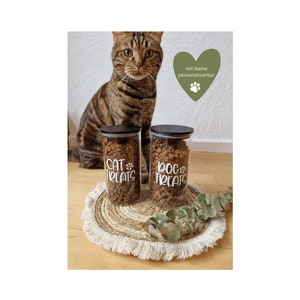 Könnte beinhalten: Zwei transparente Glasbehälter mit schwarzen Deckeln, gefüllt mit Leckerlis für Haustiere, stehen auf einer gewebten Matte. Auf einem Glas steht "CAT TREATS" und auf dem anderen "DOG TREATS" in weißer Schrift. Im Hintergrund ist eine getigerte Katze zu sehen.