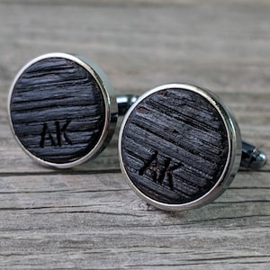 Puede incluir: Un par de gemelos de madera negra con detalles en metal plateado. Los gemelos están grabados con las iniciales "AK".