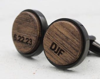May include: A pair of round cufflinks featuring dark wood inlays set in black metal frames. One cufflink is engraved with the date 6.22.23, while the other is engraved with the letters DJF in a bold, dark font across the wood grain surface.