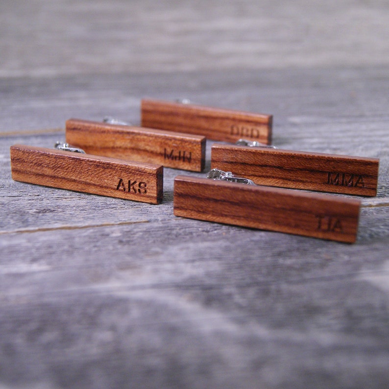 May include: Five wooden tie clips with engraved initials. The initials are "AKS", "MHN", "DBD", "MMA", and "JJA".