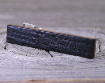 May include: A black wooden tie bar with a silver clasp. The tie bar has a natural wood grain pattern.