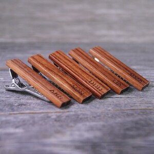 May include: Five wooden tie clips with engraved names: AIKS,  MOHN,  DIID,  TUA, and  KMNA. The tie clips are arranged in a row on a wooden surface.