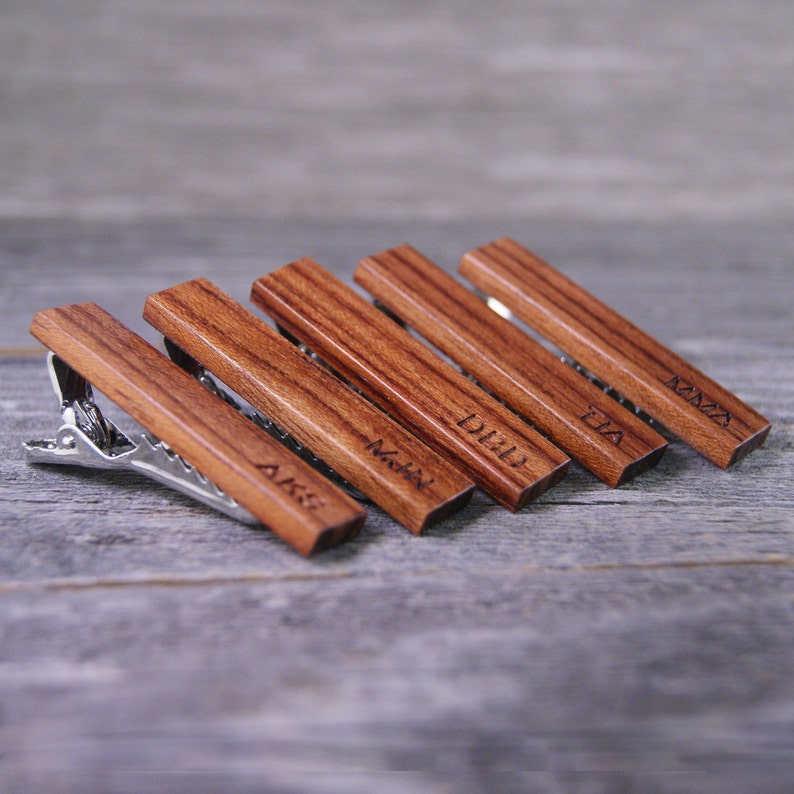 May include: Five wooden tie clips with engraved names: AIKS,  MOHN,  DIID,  TUA, and  KMNA. The tie clips are arranged in a row on a wooden surface.