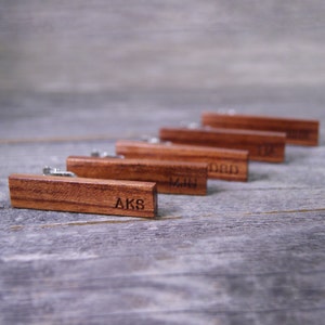 May include: A set of six wooden tie clips with engraved initials. The tie clips are arranged in a row on a gray wooden surface.