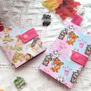 May include: Two fabric wallets with pink accents and wrist straps. One wallet features princess illustrations, the other has cartoon cat characters. Both have a snap closure and a silver-colored clasp.
