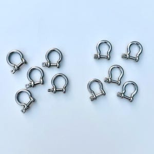 Könnte beinhalten: Eine Ansammlung kleiner, silberfarbener Schäkel in D-Form, jeweils mit Schraubverschluss. Die Schäkel sind auf weißem Hintergrund angeordnet und zeigen ihre gebogene Form und ihren metallischen Glanz. Diese Hardware-Komponenten werden häufig in verschiedenen Bastel- und Industrieanwendungen verwendet.