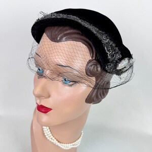 May include: A black velvet hat with a delicate net veil. The hat is adorned with silver-toned beads and a pearlescent embellishment. The hat is displayed on a mannequin head with blue eyes and red lipstick, and a pearl necklace.