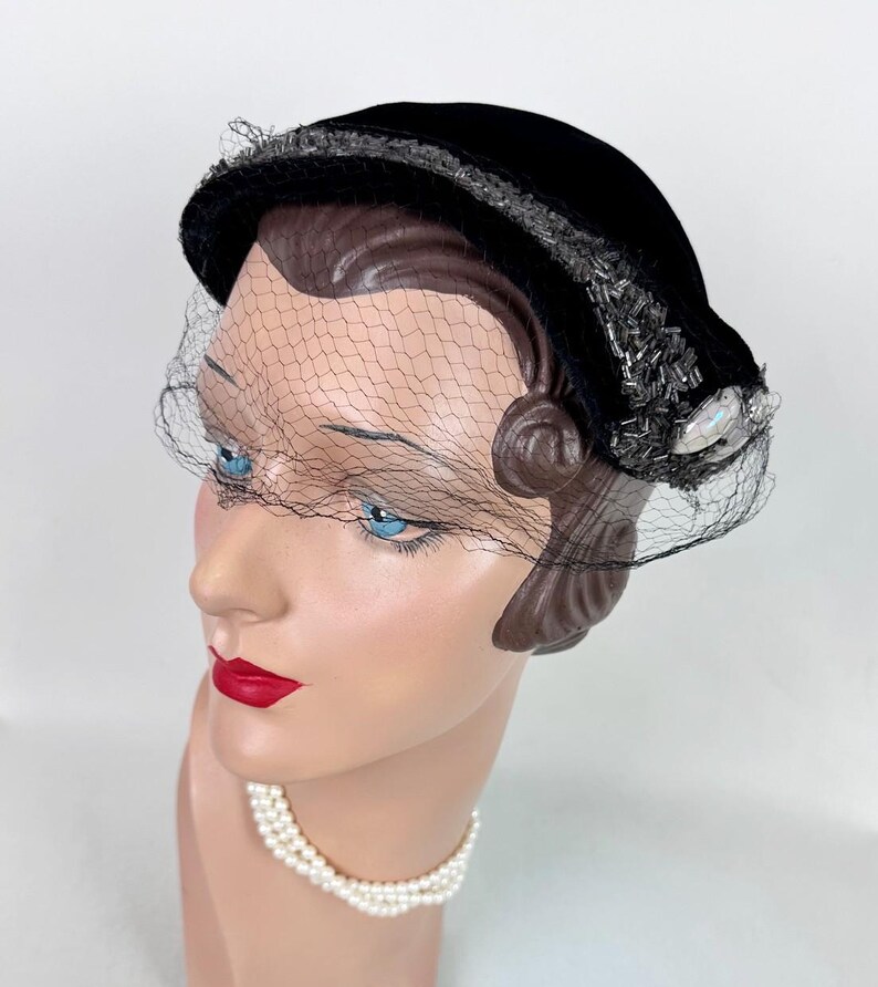 May include: A black velvet hat with a delicate net veil. The hat is adorned with silver-toned beads and a pearlescent embellishment. The hat is displayed on a mannequin head with blue eyes and red lipstick, and a pearl necklace.