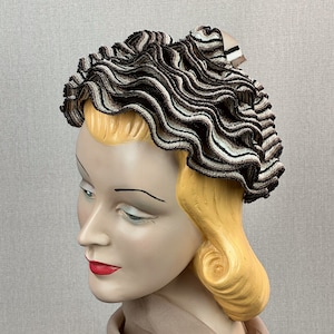 Puede incluir: Un sombrero de tela a rayas marrones y blancas con un diseño de volantes. El sombrero se lleva en un maniquí con cabello rubio.