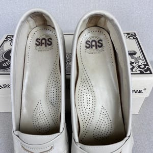 May include: A pair of white leather SAS comfort shoes, viewed from the inside. The insoles are white with small perforations. The SAS logo is visible on the insole. The shoes are in front of a white box with black text.