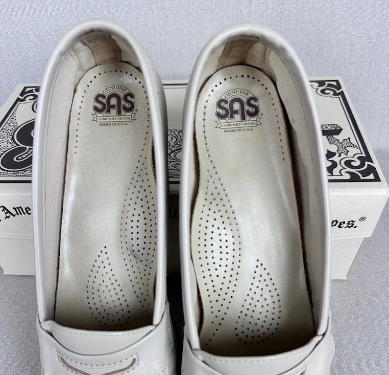 May include: A pair of white leather SAS comfort shoes, viewed from the inside. The insoles are white with small perforations. The SAS logo is visible on the insole. The shoes are in front of a white box with black text.