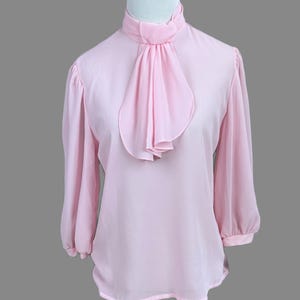 May include: Pale pink blouse with a high, ruffled collar and long sleeves. The blouse has a flowing, lightweight fabric and a soft, feminine design. The sleeves are gathered at the cuffs, adding a touch of elegance to the garment.