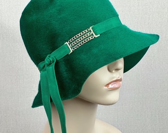 May include: A vibrant green cloche hat with a soft, textured appearance. The hat features a matching green band adorned with a silver chain detail and a bow with flowing ribbons. A stylish accessory.