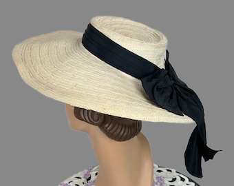Puede incluir: Un sombrero de paja de ala ancha en color beige natural con una copa plana y un tejido texturizado. Una cinta negra sólida rodea la base de la copa, atada en un lazo grande y prominente en el lateral con dos extremos largos y colgantes.