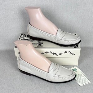 May include: Off-white leather loafers with a classic design, displayed on a shoe form. The shoes have a rounded toe, a strap across the top, and a dark brown sole. The shoes are on a box with text.
