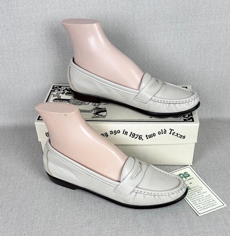 May include: Off-white leather loafers with a classic design, displayed on a shoe form. The shoes have a rounded toe, a strap across the top, and a dark brown sole. The shoes are on a box with text.