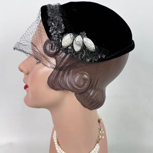 May include: A black velvet hat with a decorative beaded trim and three oval-shaped embellishments. A sheer black veil drapes over the front. The hat is displayed on a mannequin head, showcasing a vintage style.