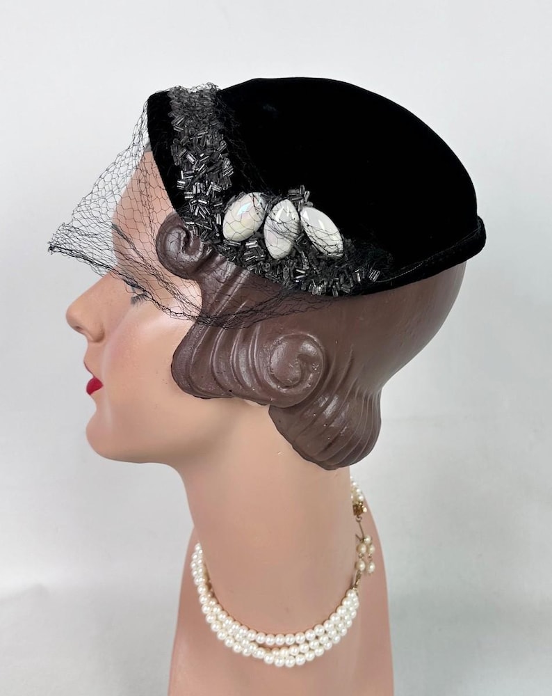 May include: A black velvet hat with a decorative beaded trim and three oval-shaped embellishments. A sheer black veil drapes over the front. The hat is displayed on a mannequin head, showcasing a vintage style.