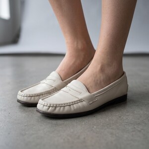 May include: Cream-coloured leather loafers with a traditional penny loafer style. The shoes feature a rounded toe, stitched vamp, and a dark brown sole. The image shows the shoes on a grey surface.