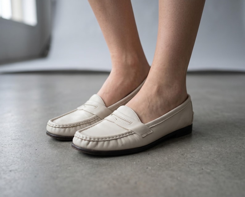 May include: Cream-coloured leather loafers with a traditional penny loafer style. The shoes feature a rounded toe, stitched vamp, and a dark brown sole. The image shows the shoes on a grey surface.